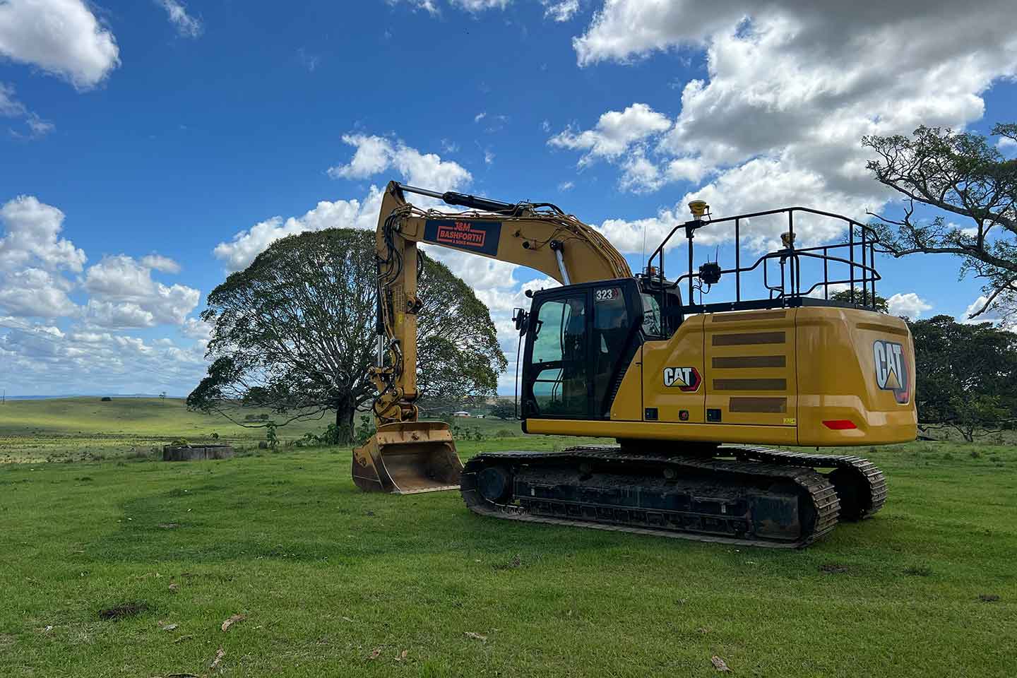 Excavator sitting on green grass in front of a large tree with a large blue sky with some clouds