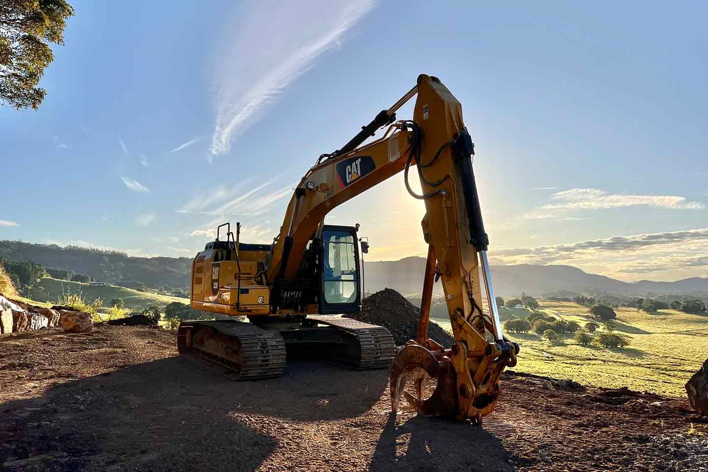 CAT excavator with grapple attachment parked on a hilltop worksite at sunrise, overlooking rolling green hills
