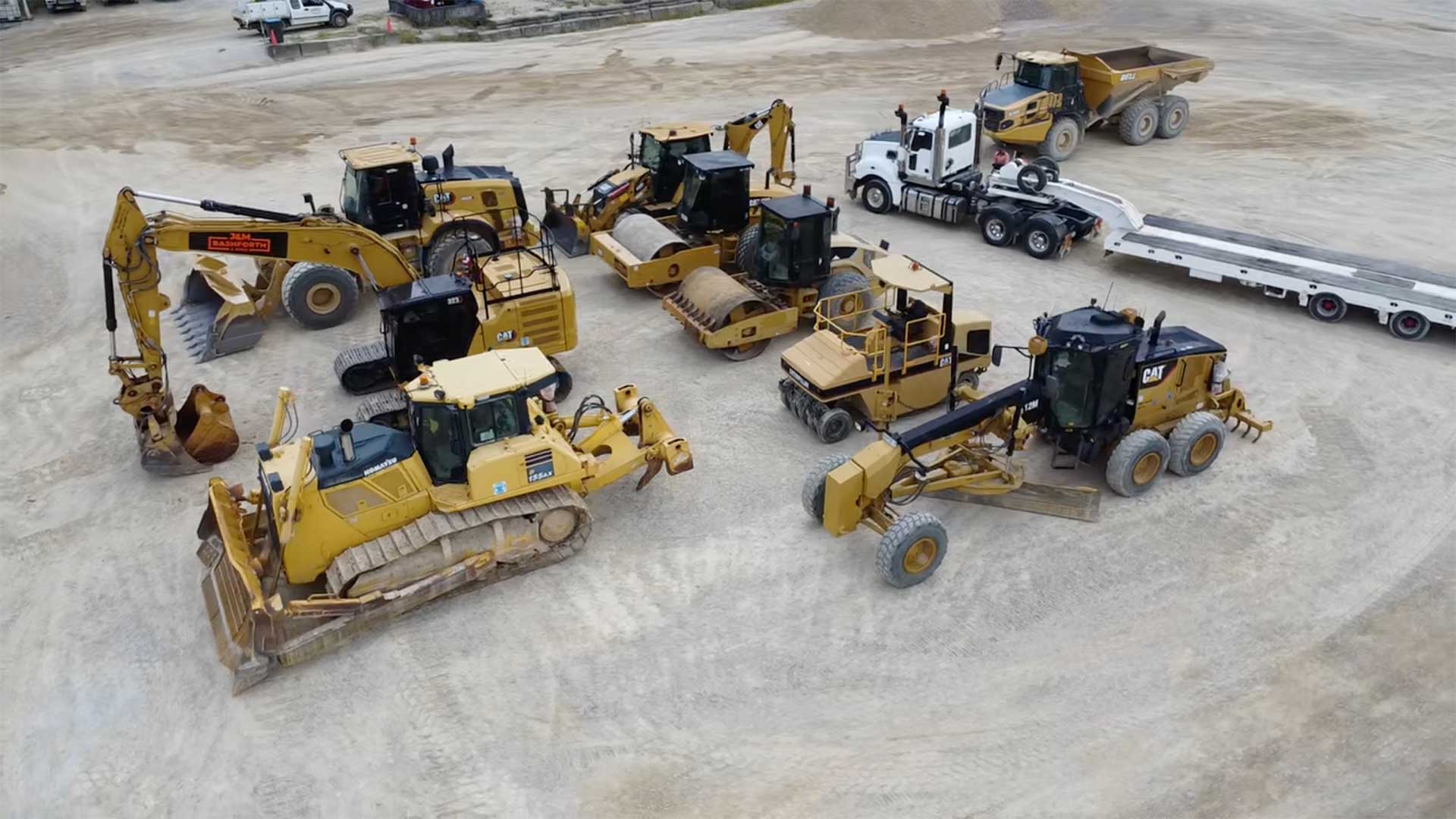 Fleet of yellow CAT and Komatsu earthmoving machinery — excavator, dozer, rollers and grader — parked with a prime mover and float trailer at a plant hire yard