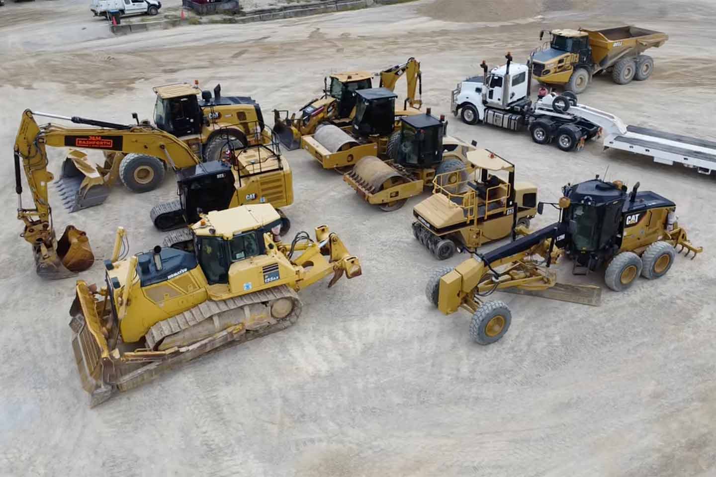 Fleet of yellow CAT and Komatsu earthmoving machinery — excavator, dozer, rollers and grader — parked with a prime mover and float trailer at a plant hire yard