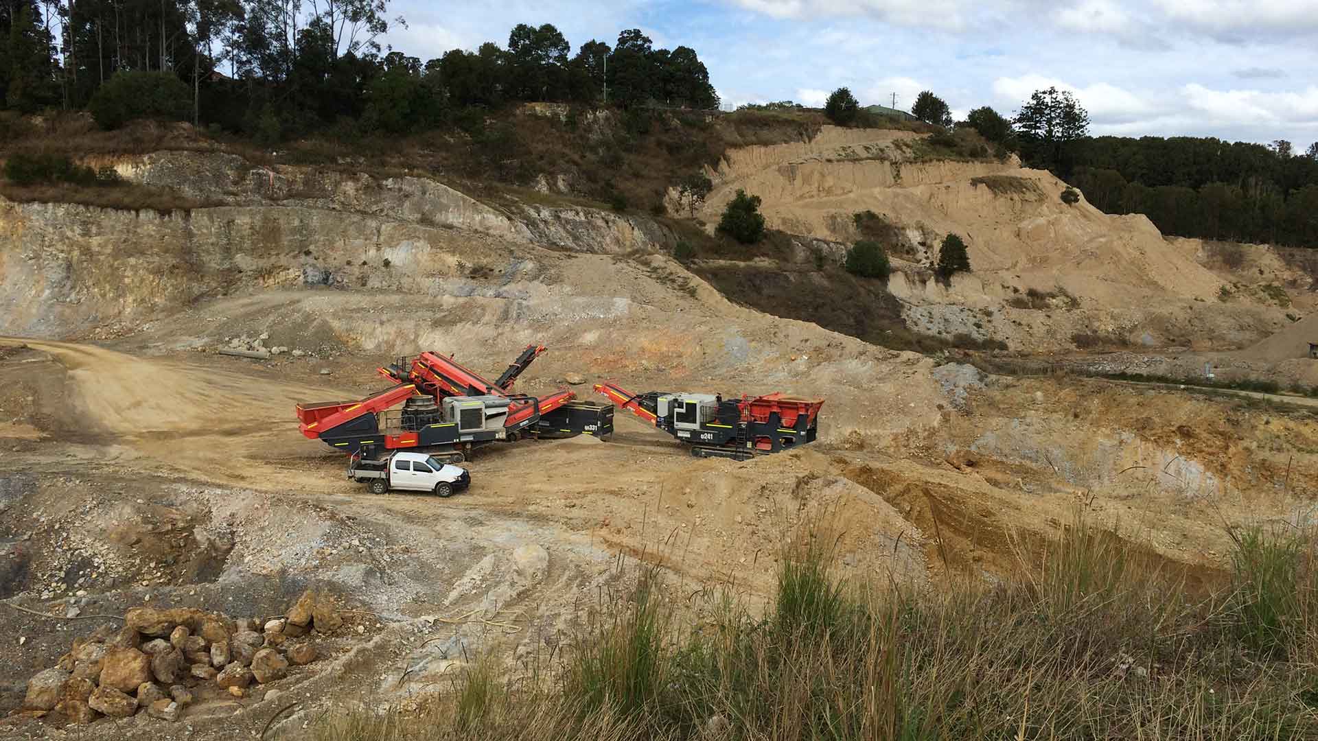 Mobile crushing and screening plant set up in an open-cut quarry, with excavated cliff faces and a ute parked alongside