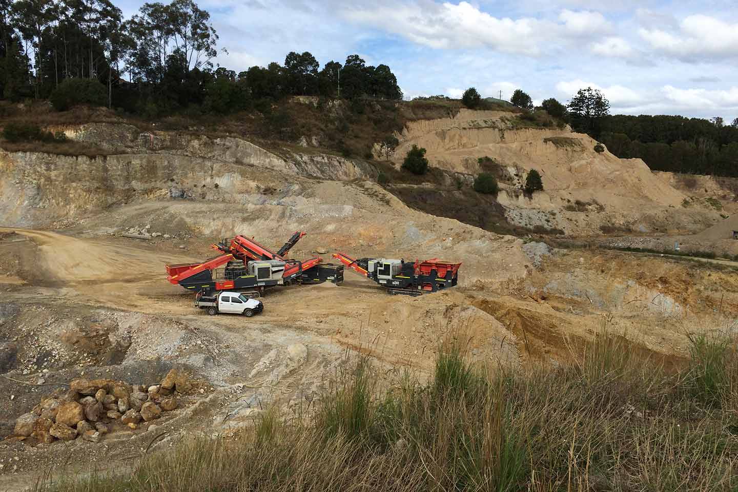 Mobile crushing and screening plant set up in an open-cut quarry, with excavated cliff faces and a ute parked alongside