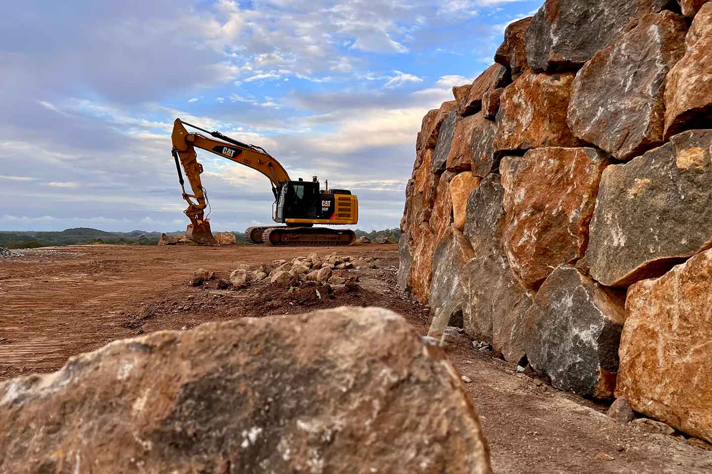 Large rock retaining wall under construction, with an excavator working on the cleared site behind it