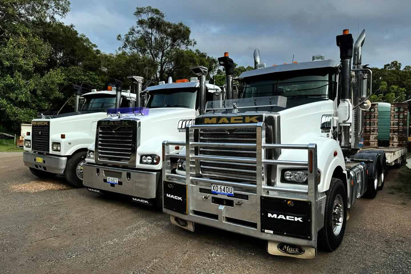 Three white Mack prime movers lined up nose-to-tail, with a flatbed trailer loaded with crates in the background